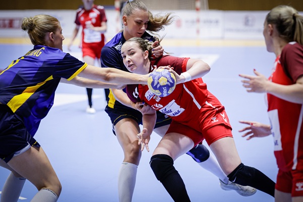 A player in a red uniform holds a handball while being closely guarded by two opponents in purple and yellow uniforms during an indoor game.